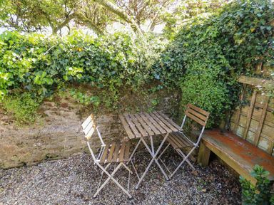 An outdoor seating area with two wooden chairs and a table on gravel near a stone wall covered with ivy and a wooden bench