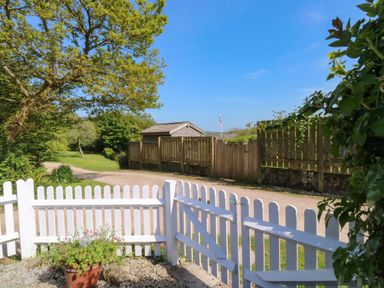 A white wooden garden gate opening to a gravel driveway with trees and a wooden fence in the background at Toad Hall Cottage in Hutcherleigh near Blackawton
