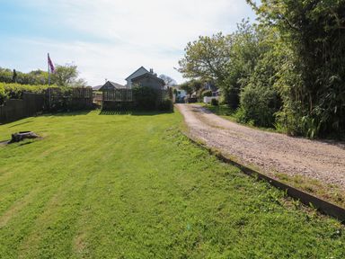 A gravel driveway beside a grassy lawn with fences trees and houses in the background at Toad Hall Cottage in Hutcherleigh near Blackawton