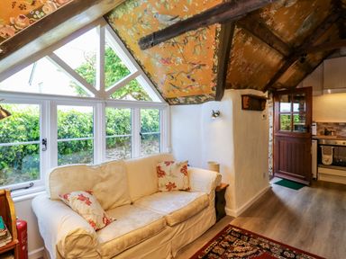 A room with a beige sofa and floral cushions by a window with patterned orange ceiling and wooden beams at Toad Hall Cottage in Hutcherleigh near Blackawton