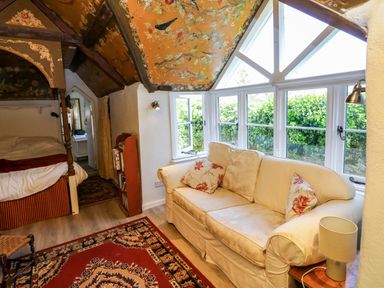 A room with a cream sofa, decorative pillows, a small lamp on a wooden side table, a patterned rug, a four-poster bed, and a large window with white frames at Toad Hall Cottage in Hutcherleigh near Blackawton