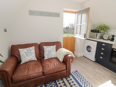 A living area with a brown leather sofa with cushions next to an open door leading outside and a kitchen area with appliances at Coach House Mews in Alderminster