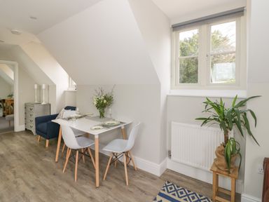 A dining area with a white table set for four and chairs under a sloped ceiling with a window and green plant on a small wooden stand at Coach House Mews in Alderminster