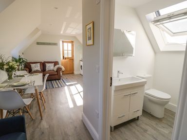 A dining area with a table set for four and a brown sofa next to a wooden door with a bathroom visible through an open door with a sink and toilet at Coach House Mews in Alderminster