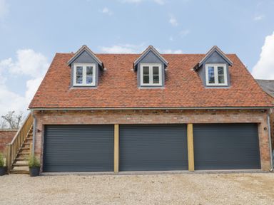 A brick building with three black garage doors three dormer windows and stairs on the side at Coach House Mews in Alderminster
