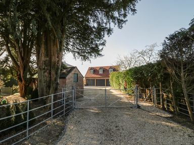 A gravel driveway with a metal gate leading to a garage with three doors surrounded by trees at Coach House Mews in Alderminster
