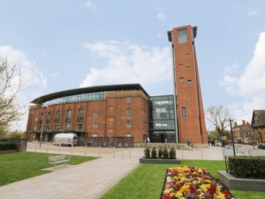 A brick building with a tall tower and glass entrance with welcome sign and flower bed in front at Coach House Mews in Alderminster