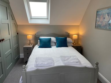 A bedroom with a bed, two blue pillows, two bedside tables with lamps, a skylight window, and a painting on the wall at Coach House Mews in Alderminster