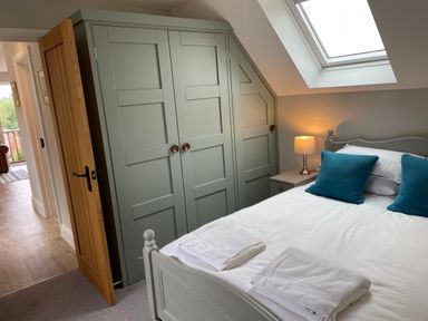 A bedroom with a bed with blue pillows white towels bedside table with lamp and built in wardrobes under a skylight window at Coach House Mews in Alderminster