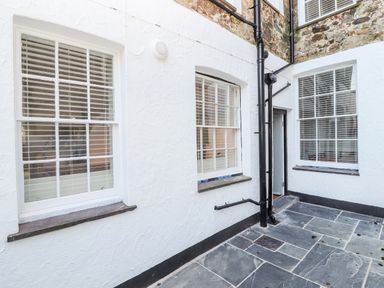 An outdoor stone patio area with three windows and a black drainpipe on a white textured wall at Lluest in Beaumaris
