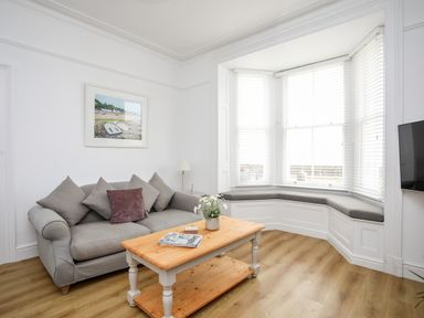A living room with a grey sofa, a wooden coffee table, a bay window with cushions, and a wall-mounted TV at Lluest in Beaumaris