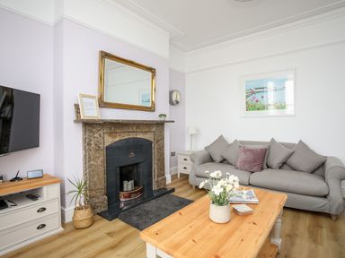 A living room with a gray sofa and cushions a fireplace with a mirror above it a wooden coffee table with flowers a television and wooden flooring at Lluest Beaumaris