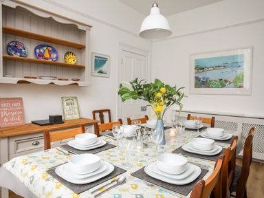 A dining room with a table set for six with white dishes and glasses a vase with yellow flowers a wall cabinet with plates and a sideboard with decorative signs at Lluest Beaumaris