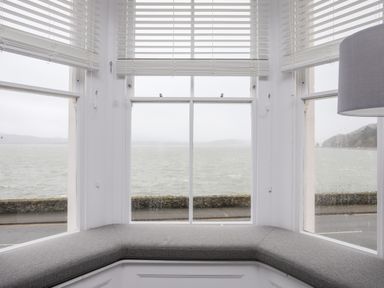 A bay window with gray cushioned seating and white blinds overlooking a body of water at Lluest in Beaumaris
