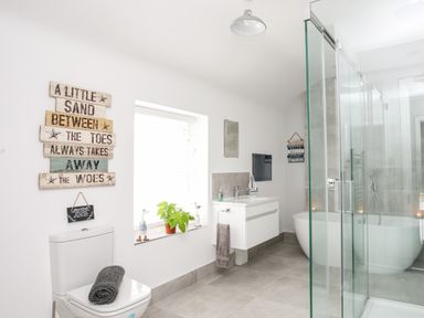A bathroom with a toilet, window with plants, wall signs, a floating sink, towel, bathtub, and glass shower enclosure at Lluest in Beaumaris