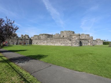 A grassy park with a paved pathway next to an old stone castle with a playground and benches at Lluest in Beaumaris