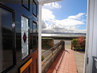 View from a black front door opening to a tiled pathway with metal fence and plants overlooking a road and water with boats and mountains in the distance at Lluest in Beaumaris