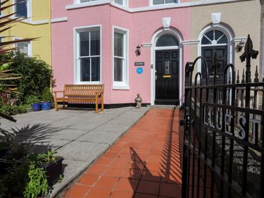 An entrance area with a black gate and a pathway leading to black doors of a pastel pink and beige building with a wooden bench and plants at Lluest in Beaumaris