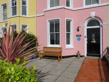 A pink and yellow house exterior with a black door wooden bench and plants at Lluest in Beaumaris