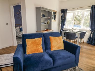 A living area with a blue sofa with two orange cushions a dining table with four chairs and a grey cabinet at Primrose End in Jacobstow