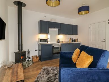A kitchen area with navy cabinets and appliances next to a wood burning stove and a blue sofa with yellow cushions at Primrose End in Jacobstow
