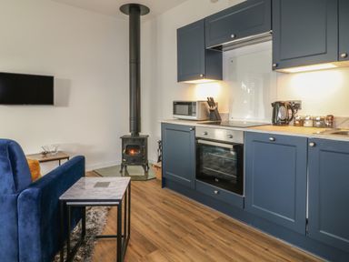 A kitchen with blue cabinets a built in oven microwave kettle a wood burning stove and a blue sofa with nested tables at Primrose End in Jacobstow