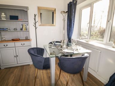 A dining area with a glass table set for four and blue chairs next to a window and a gray cabinet at Primrose End in Jacobstow