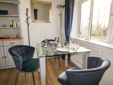 A dining area with a glass table set for two and blue chairs near a window at Primrose End in Jacobstow