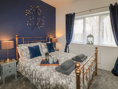 A bedroom with a bed with patterned bedding and blue pillows and curtains with bedside tables and lamps and a window at Primrose End in Jacobstow