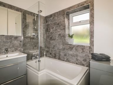 A bathroom with a bathtub shower combination a sink cabinet a window with a potted plant and folded towels at Primrose End in Jacobstow