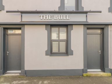 The exterior of a building with two grey doors and a window under a sign that reads THE BULL at The Bull Inn in Deiniolen