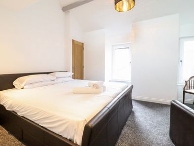 A bedroom with a large bed, white bedding, towels on the bed, a wooden door, two windows, a chair, and carpeted floor at The Bull Inn in Deiniolen