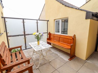 An outdoor patio area with a wooden bench, a wooden pew-style seat, a white metal table with a vase of flowers and frosted glass panels at The Bull Inn in Deiniolen