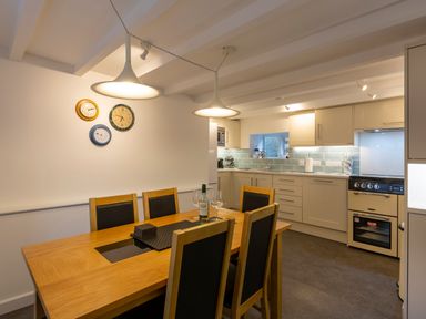 A dining area with a wooden table and six chairs beside a kitchen with light-colored cabinets and a stove at Rose Cottage in St Ives