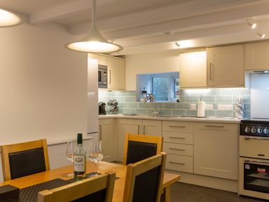 A kitchen with cream cabinets a blue tiled backsplash a wooden dining table with chairs and a bottle of wine at Rose Cottage in St Ives