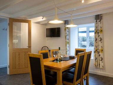 A dining room with a wooden table and six chairs with black cushions a wine bottle and glasses on the table pendant lights above and a window with floral curtains at Rose Cottage in St Ives