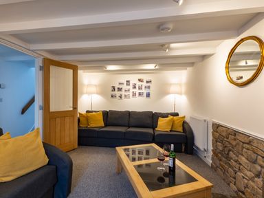 A living room with a dark sofa and yellow cushions two floor lamps a coffee table with wine glasses and a bottle a wooden door and a round mirror on the wall at Rose Cottage in St Ives
