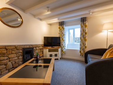 A living room with a stone wall fireplace a TV on a white cabinet a gray sofa with yellow cushions a wooden coffee table with two wine glasses and a bottle a window with floral curtains and a floor lamp at Rose Cottage in St Ives