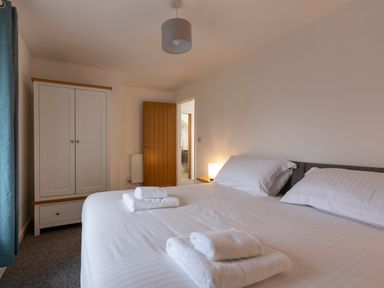 A bedroom with a double bed with white linens and towels on top a white wardrobe a wooden door a bedside table with a lamp and blue curtains at Rose Cottage in St Ives