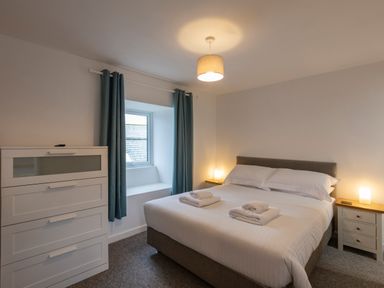 A bedroom with a double bed white linens bedside tables with lamps a chest of drawers and a window with blue curtains at Rose Cottage in St Ives