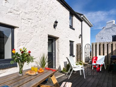 An outdoor patio with a wooden picnic table with flowers and food and white chairs next to a white stone building at Rose Cottage in St Ives
