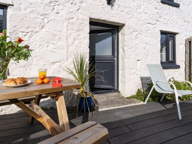 An outdoor wooden picnic table with croissants oranges flowers and a glass of juice next to a white stone building with a half open black door and a white chair on a dark deck at Rose Cottage in St Ives