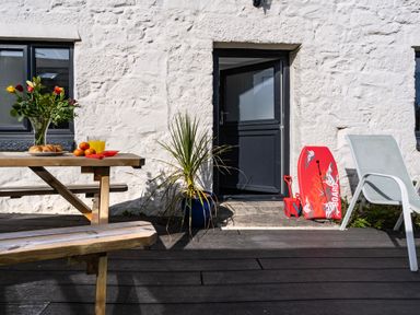 A patio with a wooden picnic table with croissants juice and flowers a plant in a pot a beach chair and a red boogie board by a black door on a white stone wall at Rose Cottage in St Ives