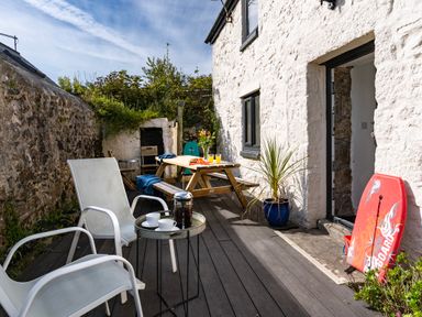 An outdoor patio with white chairs a small table with a coffee press and cups a wooden picnic table with drinks and flowers and a red bodyboard at Rose Cottage in St Ives