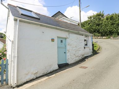 A small white cottage with a blue door and window on a sloping road at The Old Stable in Porthallow
