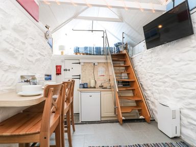 A small kitchen area with a wooden staircase leading to a loft bed and dining table with chairs in a white stone walled room at The Old Stable in Porthallow