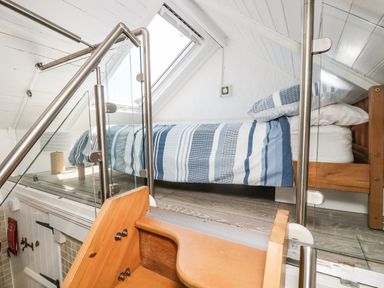 A loft bedroom with a single bed with blue striped bedding accessed by wooden stairs with glass and metal railing at The Old Stable in Porthallow
