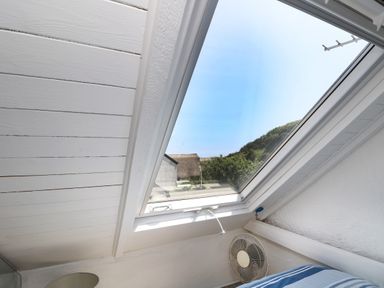 An angled skylight window showing a view outside with a fan and part of a bed inside the room at The Old Stable in Porthallow