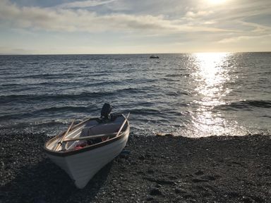 A small boat on a rocky shore with the sea and sunlight reflecting on the water at The Old Stable in Porthallow