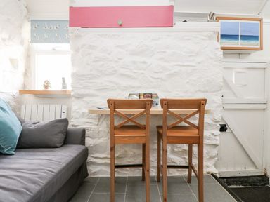 A small dining area with two wooden chairs and a table against a white stone wall near a window and a white door at The Old Stable in Porthallow